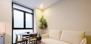 Una sala de estar moderna con un sofá, una mesa de comedor con sillas, una planta en maceta y un ventanal grande.