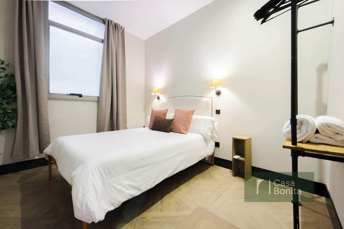 Habitación de hotel ordenada con cama, una mesita de noche, un mueble auxiliar, estante con toallas y ventana con cortinas.