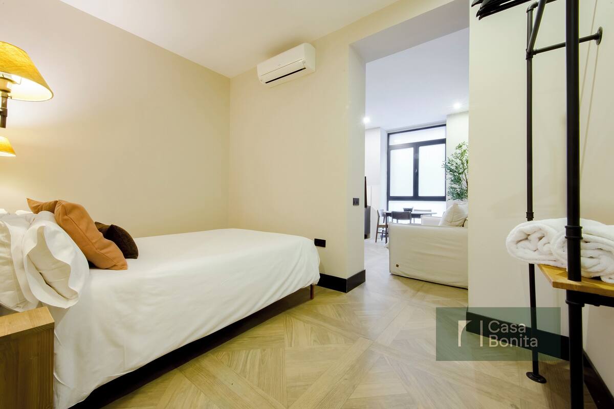 Habitación de hotel con cama, mesita de noche, unidad de aire acondicionado y vista de la habitación contigua.