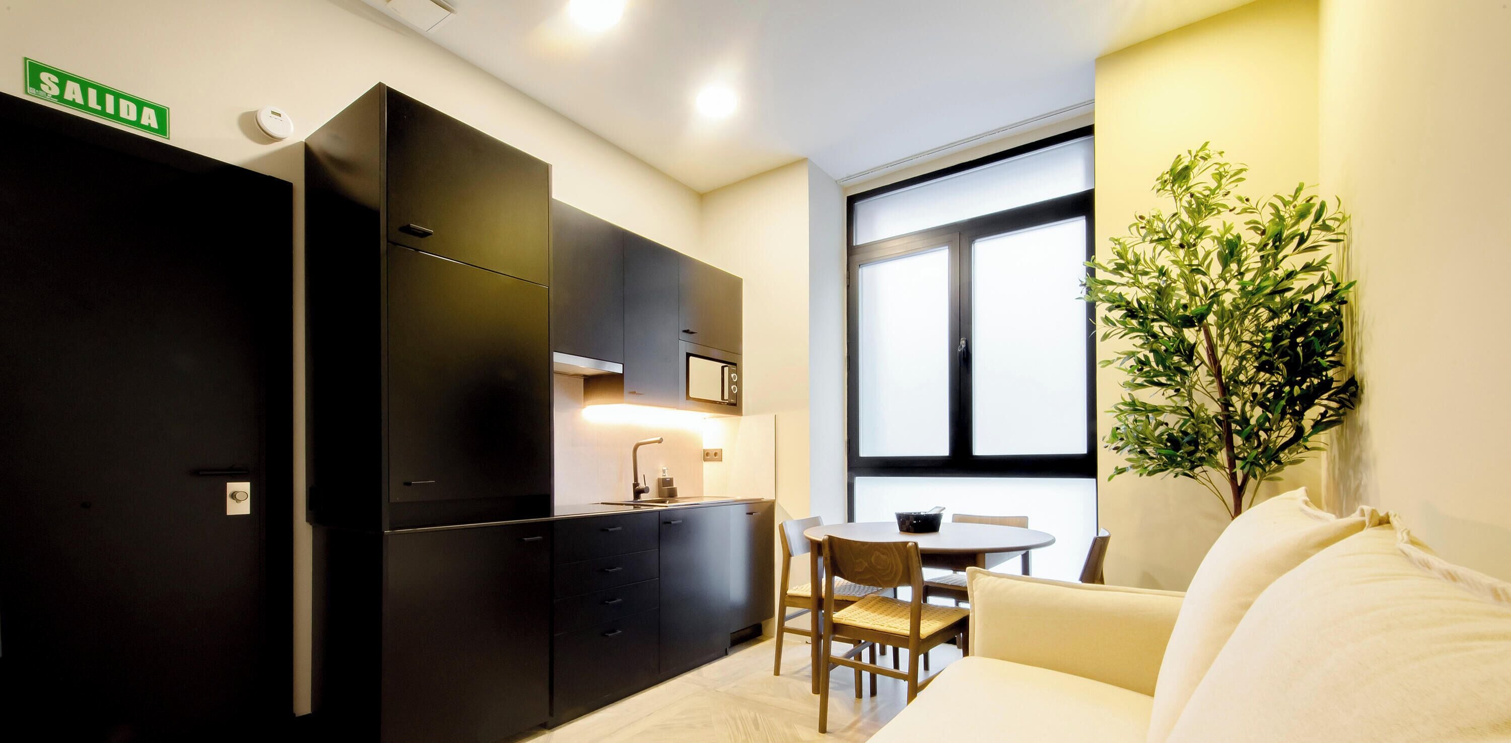 A compact kitchen with black cabinets, a dining area with a round table and chairs, a white sofa, and a large window with a view.