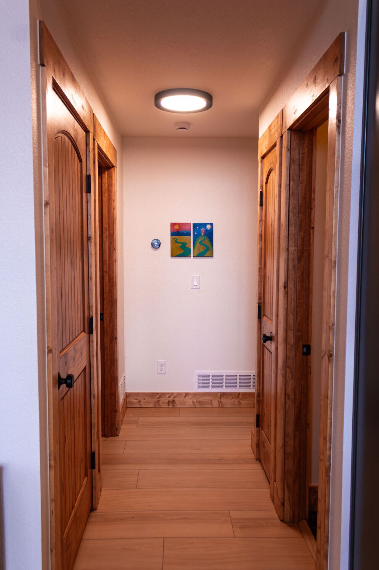 A narrow hallway with wooden doors on either side, a ceiling light, and two small wall-mounted artworks.