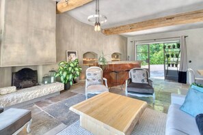 A spacious living room with a fireplace, stone flooring, and a large window offering a view of greenery. - Magnifique Maison historique (Saint Paul de Vence)