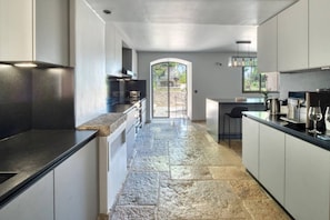 A modern kitchen with stone flooring, a central island, and a view through an arched doorway. - Magnifique Maison historique (Saint Paul de Vence)