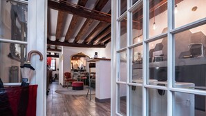 Un intérieur spacieux avec de hauts plafonds et des poutres apparentes. Une grande fenêtre donne sur une salle de bain équipée d'une baignoire et d'un espace douche. La pièce est dotée d'un plancher en bois et d'une chaise rouge placée au centre.