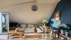 Een gezellige woonkamer met een bank, salontafel en een eettafel met stoelen. Er is een televisie op een dressoir, een decoratief kunstwerk aan de muur en een potplant.