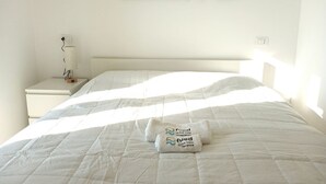A neatly made bed with a white comforter and two towels with text, a bedside table with a lamp, and a painting on the wall. - CV020 – Cervia, penthouse with stunning sea view (Cervia)
