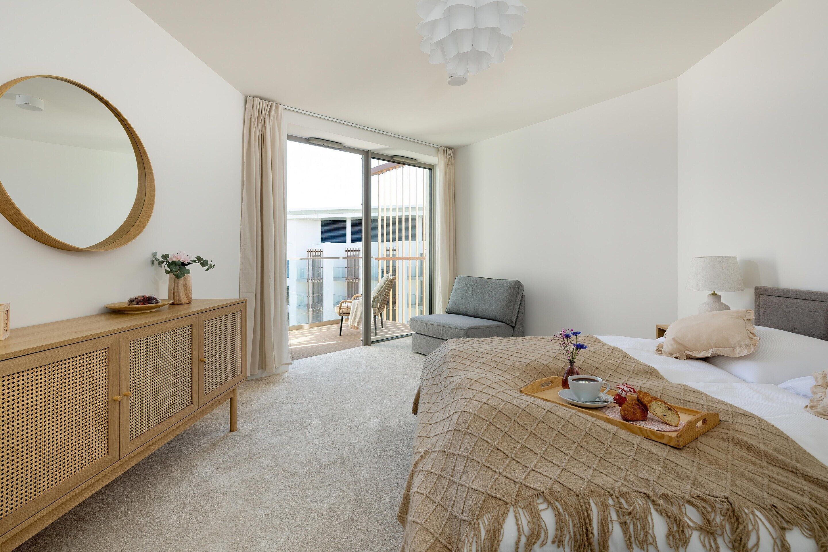 Ein Hotelzimmer mit Bett, Nachttisch, Spiegel, Kommode, einem Balkon mit Stuhl und einem Tablett mit Frühstücksgegenständen.