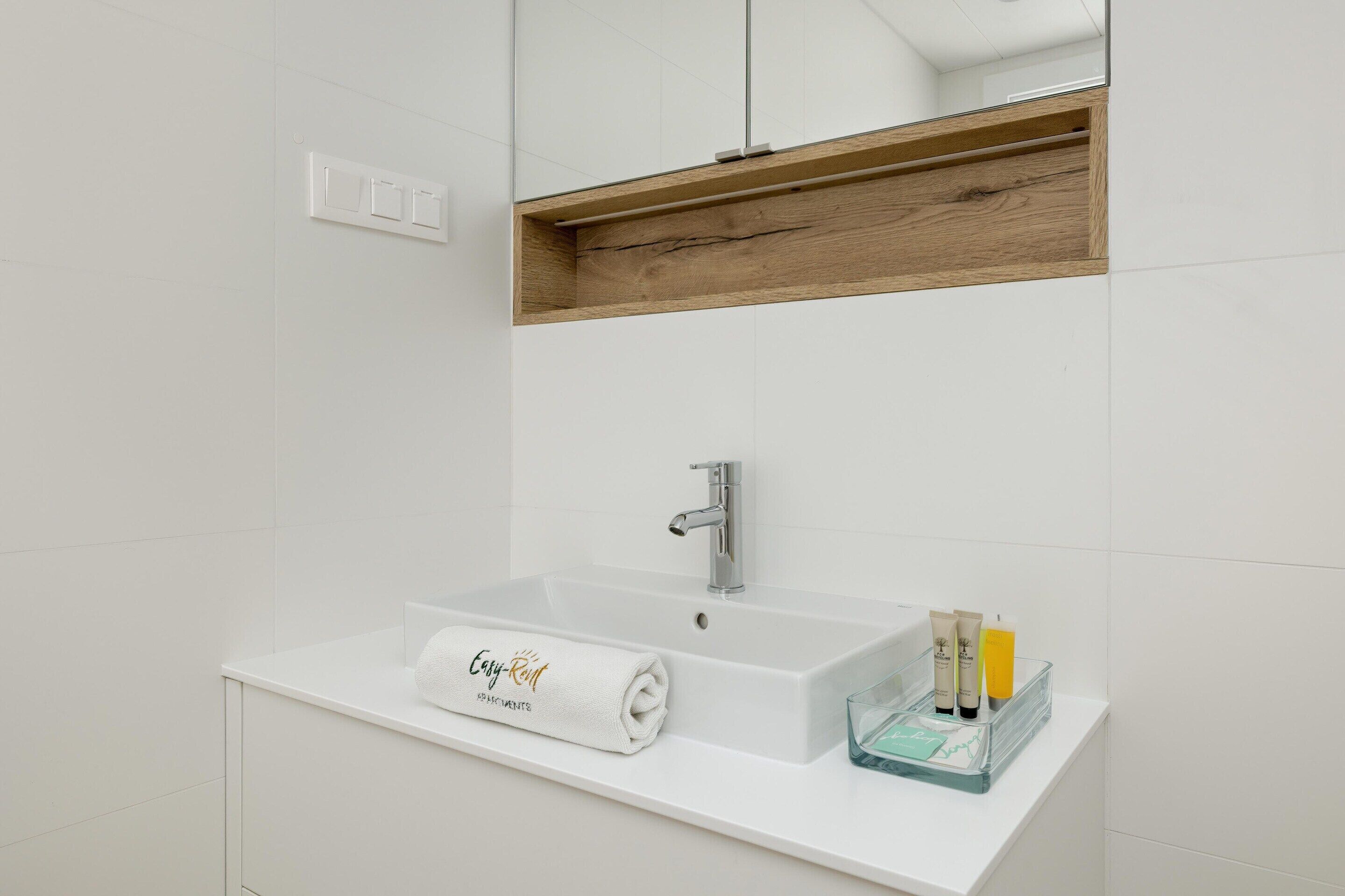 A modern bathroom with a white sink, a wooden shelf above the mirror, and a towel with "Easy Room" text.