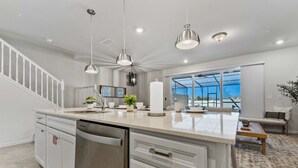 Een moderne keuken met een groot eiland, roestvrijstalen apparatuur, hanglampen en uitzicht naar buiten.