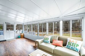 Ein Wintergarten mit großen Fenstern, Holzboden, einem beigefarbenen Sofa mit Strandkissen und einem kleinen Tisch.