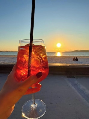 A hand holding a glass with a straw, a sunset in the background, and a beach scene. - Mediterranean life style, sea view, 10 min. walk from city centre. (Alghero)