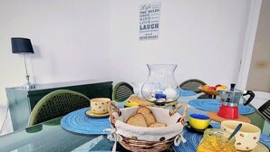 Uma mesa de jantar com tampo de vidro, rodeada por cadeiras, uma cesta de pão, uma cafeteira e uma placa na parede.