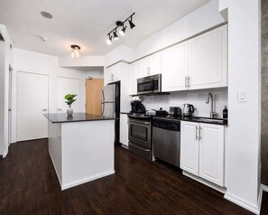 A modern kitchen with white cabinets, a central island, stainless steel appliances, and a dark wood floor. - Charming 1BR+Den in Downtown Toronto with Balcony (Toronto)