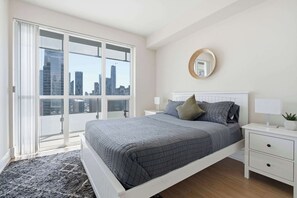A bedroom with a bed, bedside tables, a mirror, and a view of the cityscape through large windows. - Cityscape Haven 2BR with Fantastic CN Tower View (Toronto)
