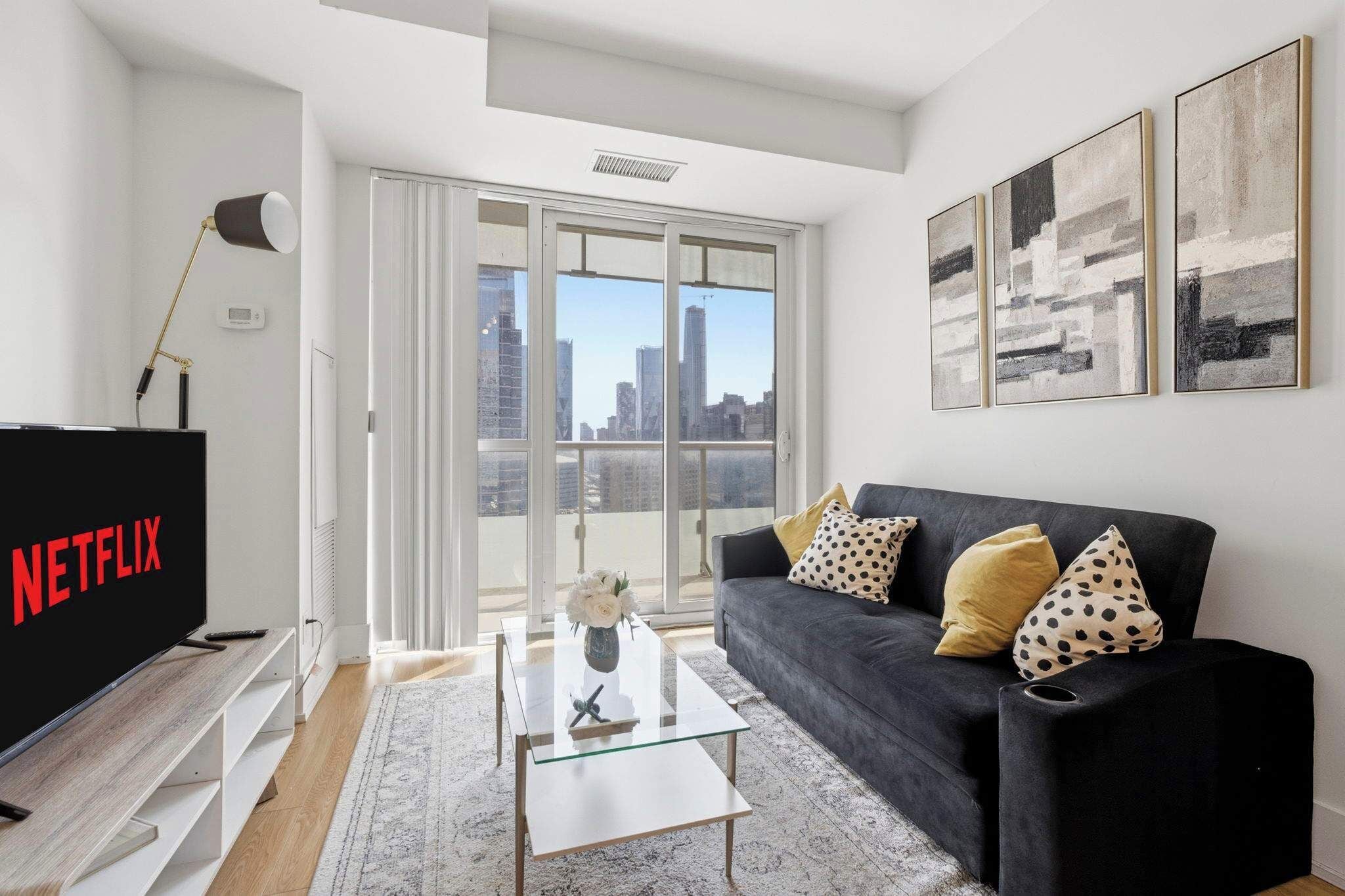 A modern living room with a black sofa, a glass coffee table, a TV with 'NETFLIX' on it, and a city view through large windows.
