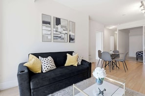A modern living room with a black sofa, a glass coffee table, and a dining area with a table and chairs. - Cityscape Haven 2BR with Fantastic CN Tower View (Toronto)