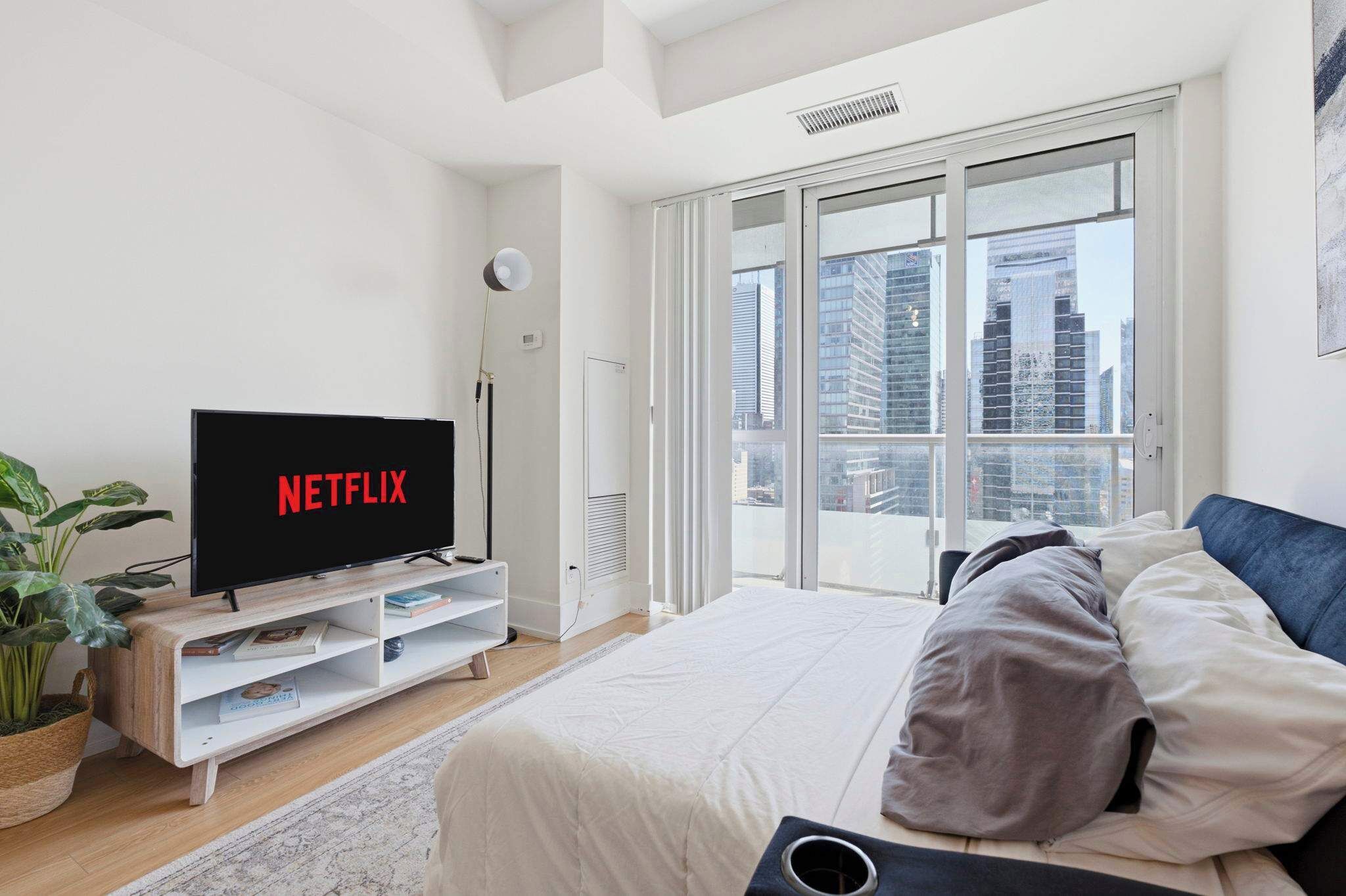 Une chambre moderne avec un grand lit, un téléviseur posé sur un meuble en bois et une vue sur la ville grâce à de grandes fenêtres.