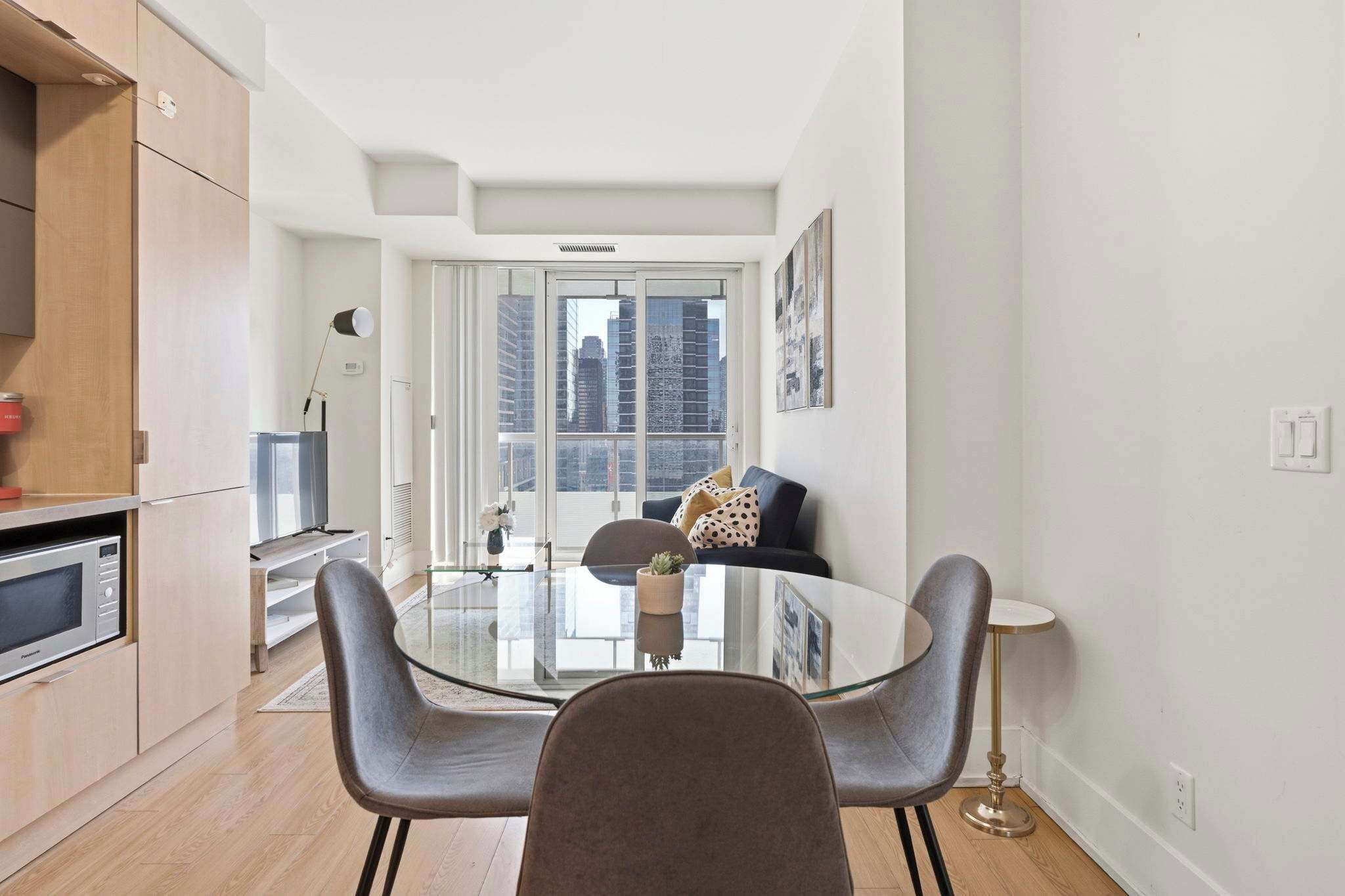 Een moderne keuken met een ingebouwde magnetron, een eethoek met een glazen tafel en stoelen, en een groot raam met uitzicht op de stad.