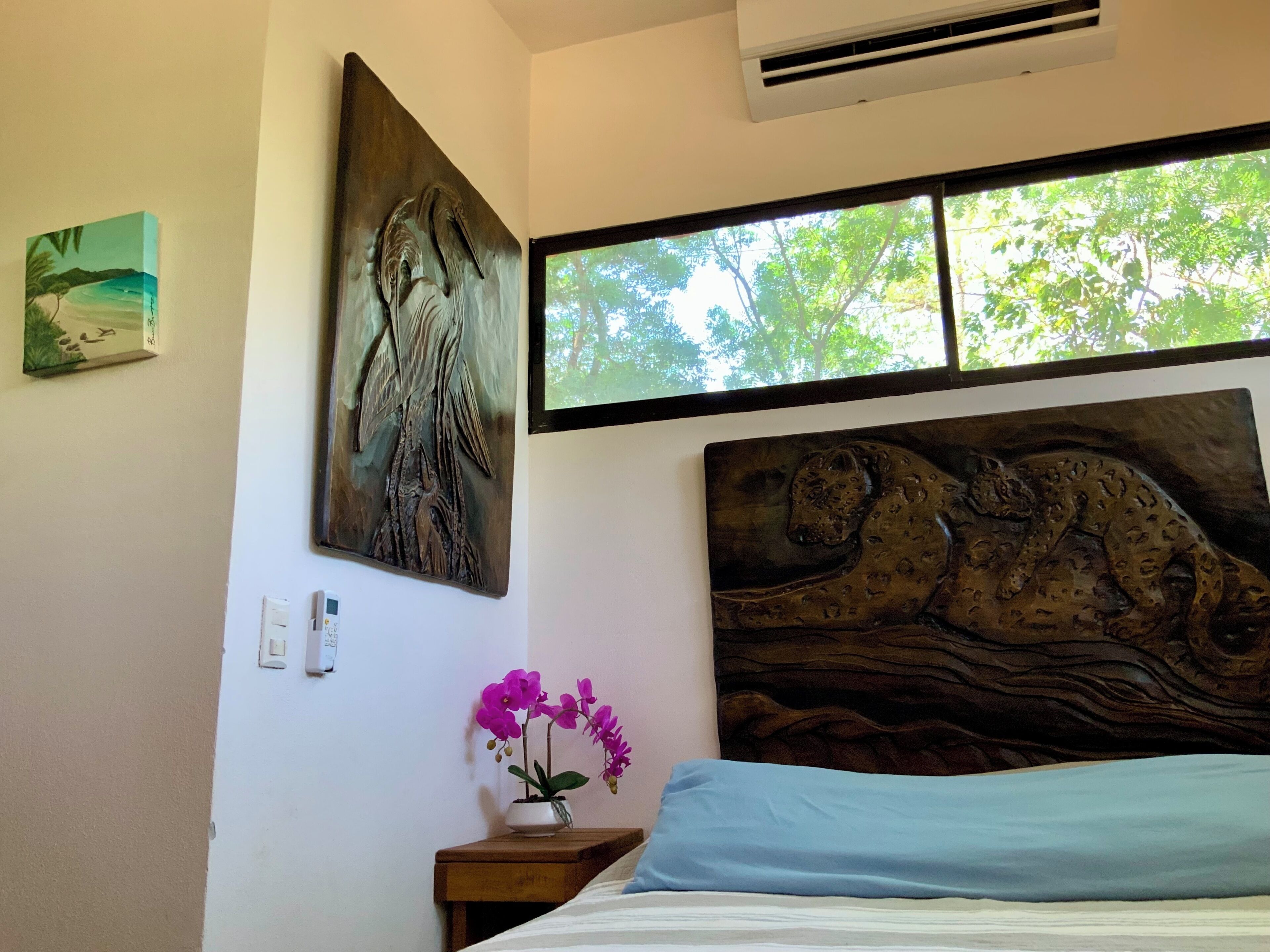Slaapkamer met een bed, een raam met uitzicht op bomen, een wandairconditioner en een schilderij van een boomstam.