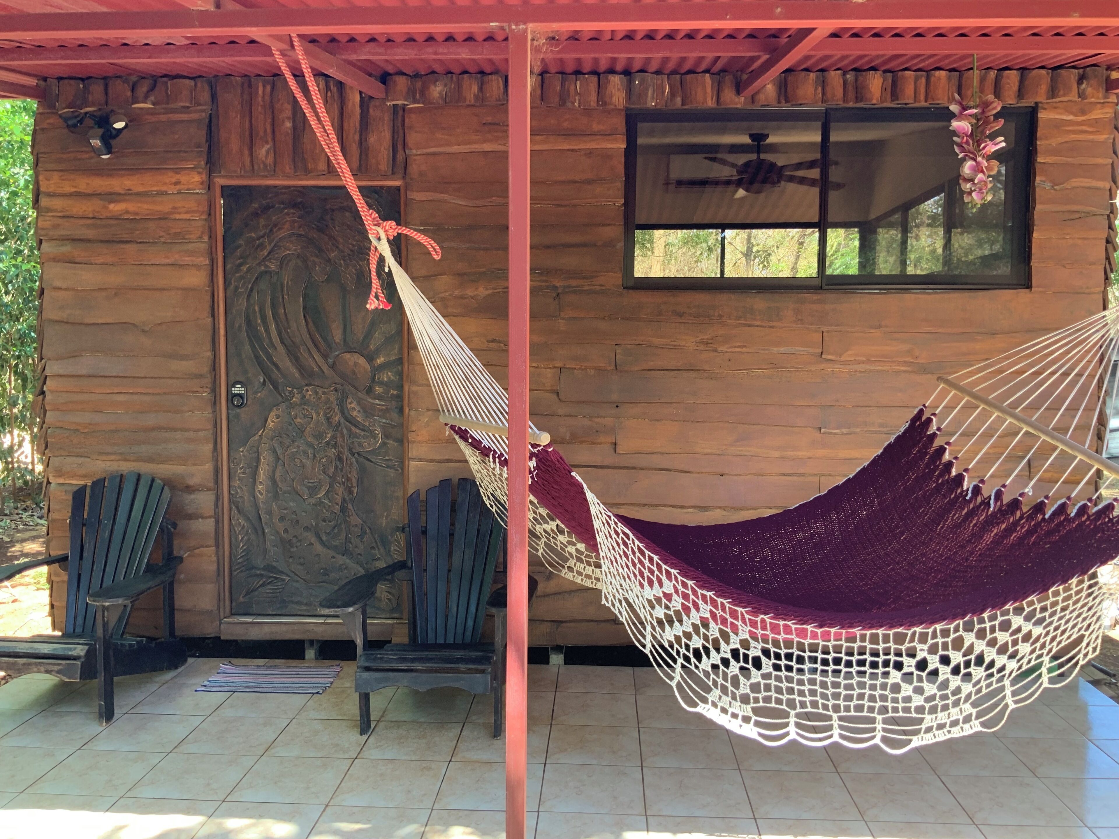 Un hamac suspendu entre deux poteaux, des chaises en bois, une porte ornée d’un panneau décoratif et une fenêtre avec un ventilateur de plafond.