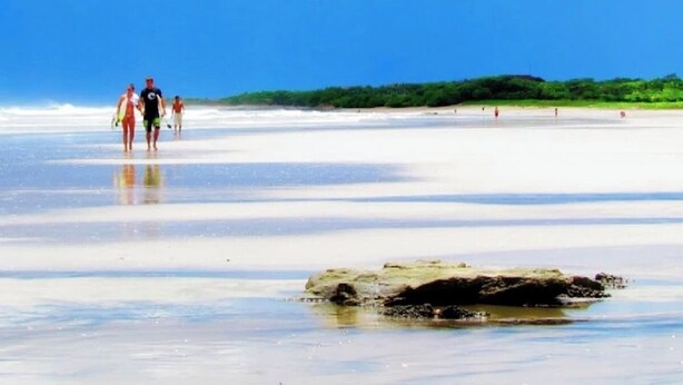 A beach with people walking, a large rock in the foreground, and a clear sky. - Playa Avellanas Hand-Carved Casita Walk to Beach Private Jungle Stay (Playa Avellanas)