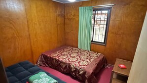 A small, dimly lit room with wooden walls, a single bed with a patterned bedspread, a small table with a red object, and a window with green curtains. - Black Stone (Licanray)