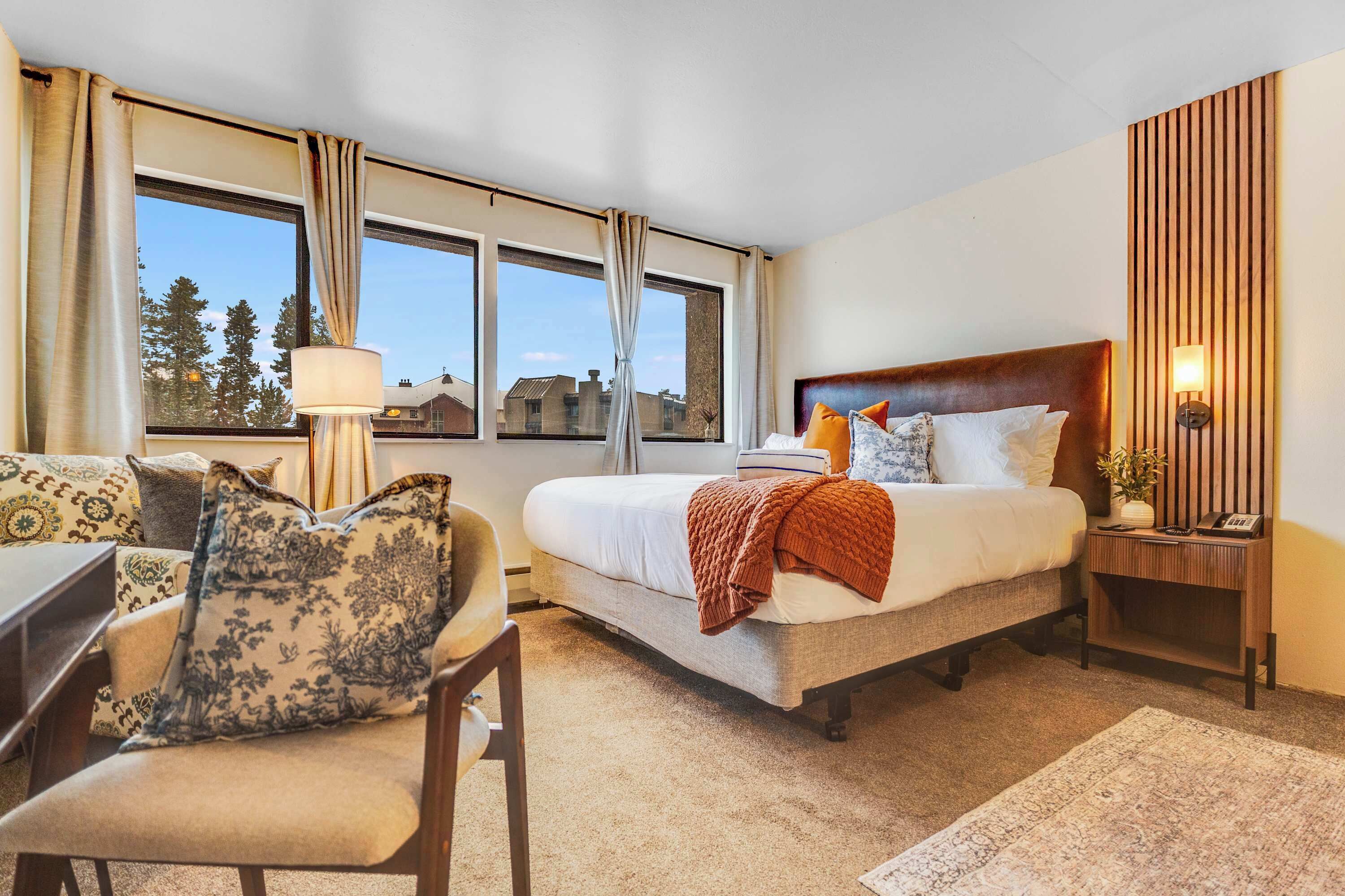 A hotel room with a bed, a desk, a chair, a lamp, a nightstand, and a view of trees and buildings through the window.