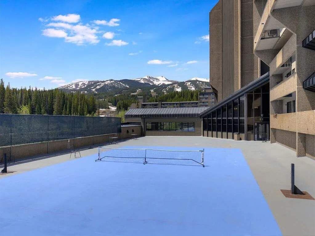 A tennis court with a mountain backdrop, adjacent to a modern building with large windows and balconies.