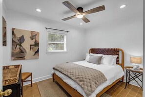 A bedroom with a wooden bed, a ceiling fan, a window with blinds, and a painting on the wall. - Downtown Magnolia (Ocean Springs)