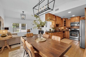 A modern kitchen with wooden cabinets, a dining table with chairs, a chandelier, and a large window. - Downtown Magnolia (Ocean Springs)