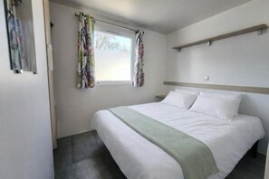 Ein Schlafzimmer mit einem Bett, einem Fenster mit Vorhängen, einem Nachttisch und einem Kopfteil.