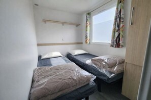 Eine kleine, einfache Unterkunft mit zwei Betten, einem mit Vorhängen versehenen Fenster und einem Holzregal über den Betten.