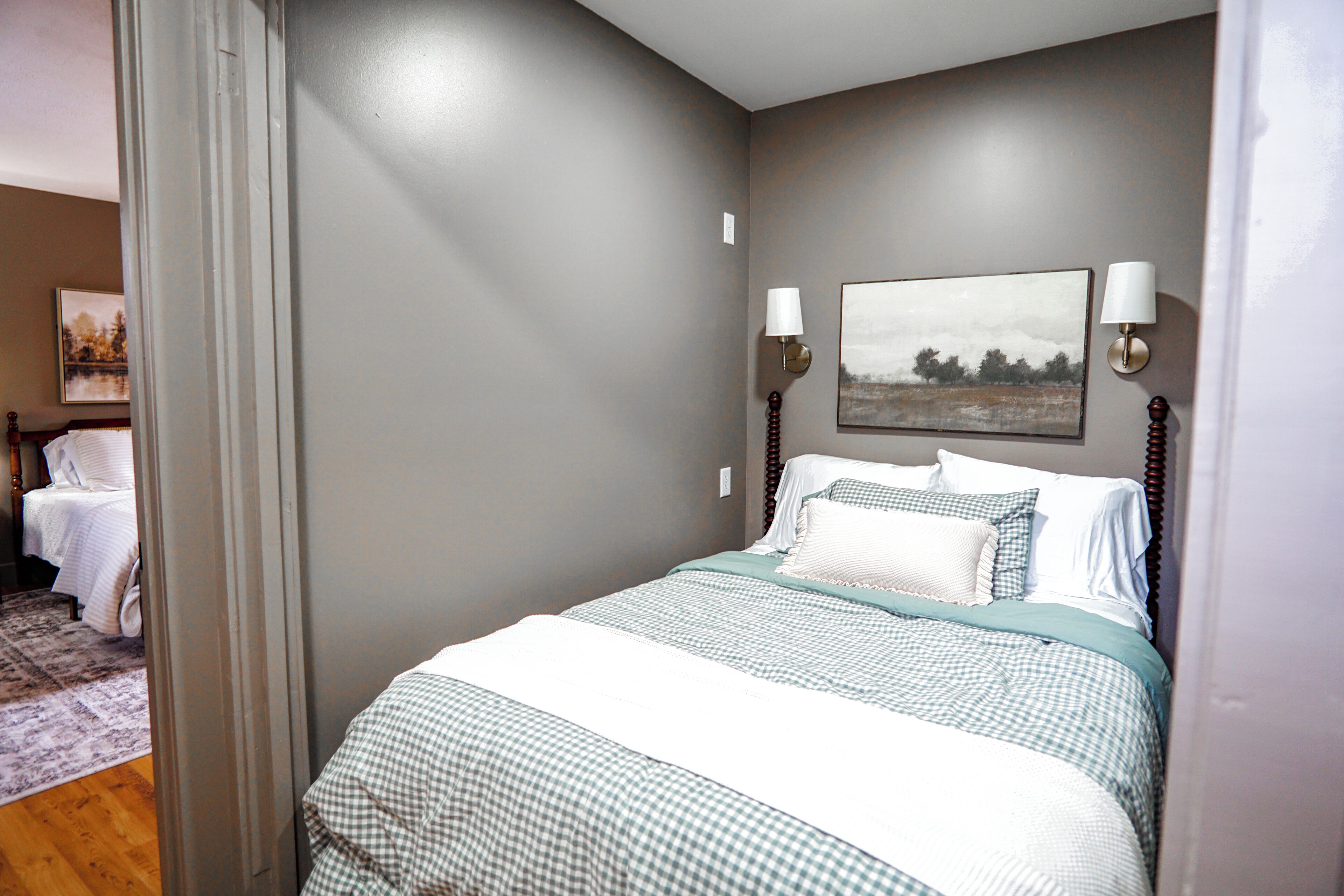 A neatly made bed with checkered bedding, two wall-mounted lamps, and a framed landscape painting above the bed.