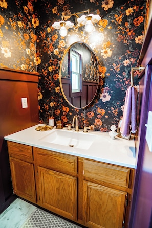 A bathroom with a floral wallpaper, a round mirror, a wooden vanity, and a purple towel hanging on the wall.