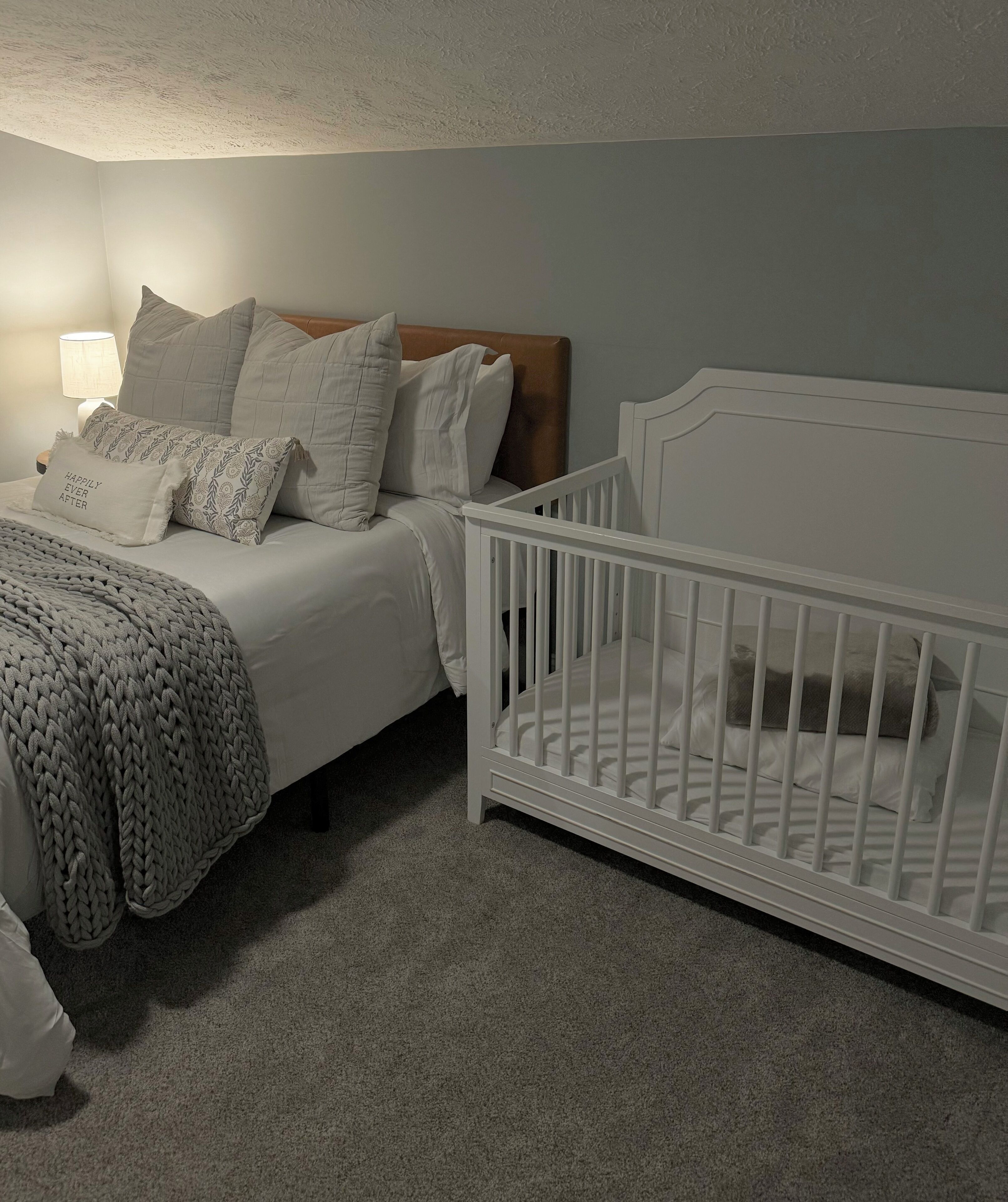 A bedroom with a bed, a crib, a lamp, and a textured throw blanket.