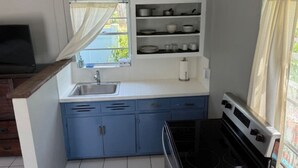 Uma cozinha compacta com armários azuis, bancada branca, fogão e uma janela com cortinas.