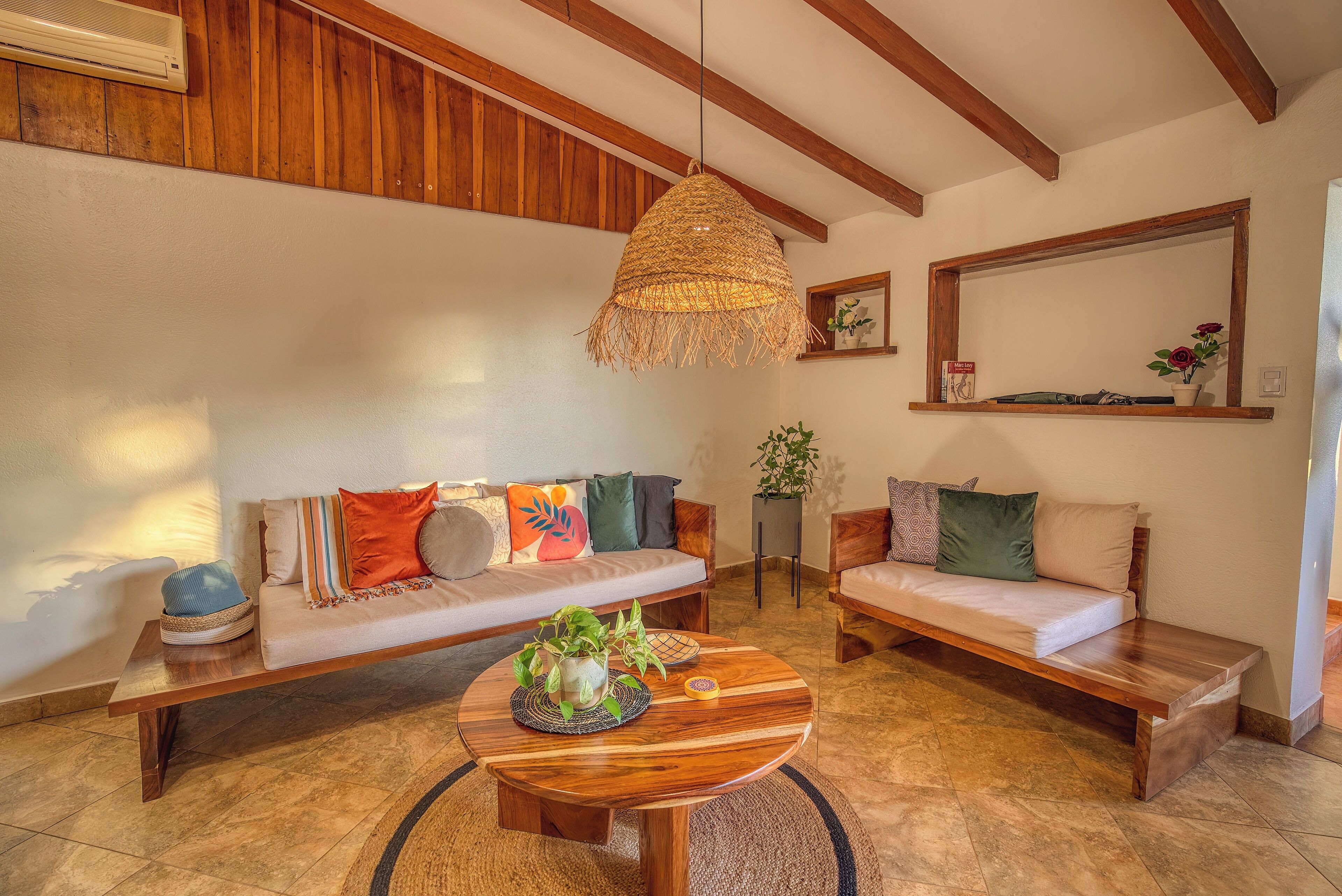 A cozy living room with a wooden bench, a round coffee table with a woven hanging light, and a sofa with colorful cushions.