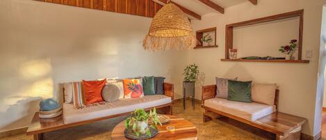 A cozy living room with a wooden bench, a round coffee table with a woven hanging light, and a sofa with colorful cushions.