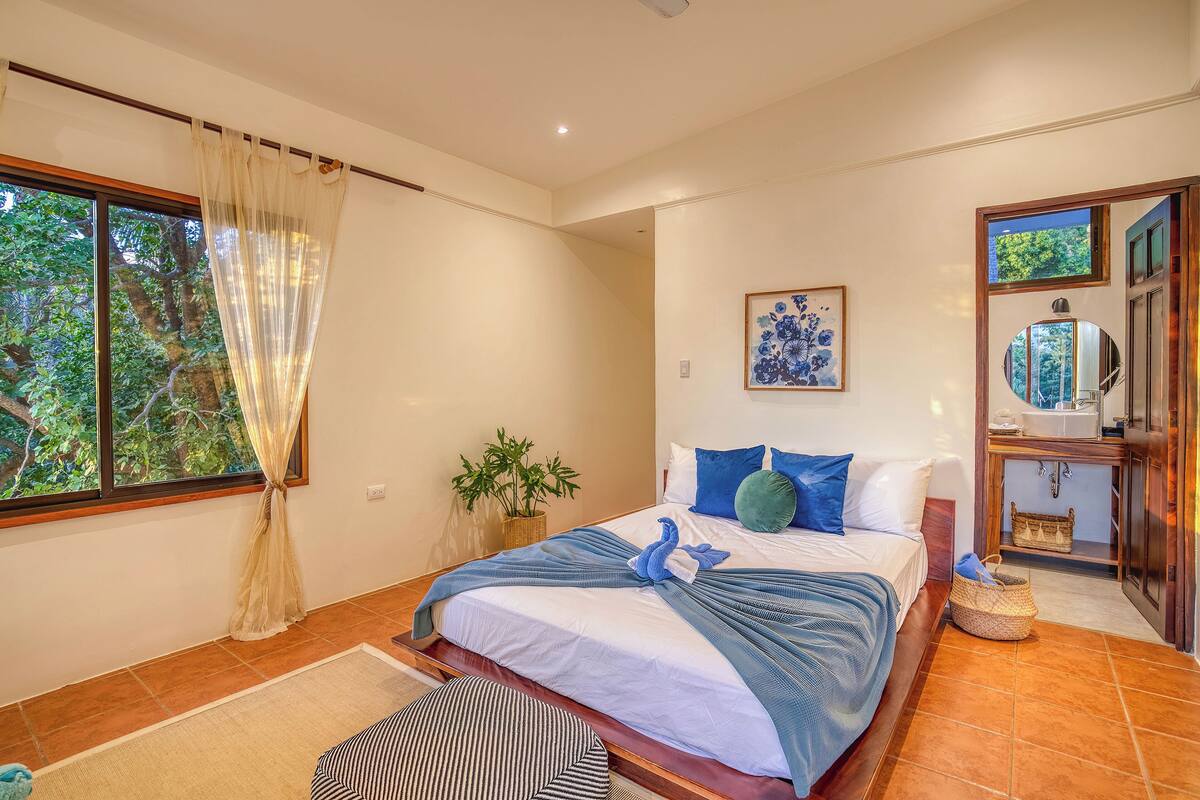 A bedroom with a large bed, a window with curtains, a potted plant, and a wall-mounted artwork.
