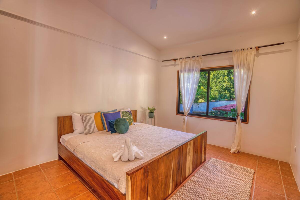 A bedroom with a wooden bed, a view of greenery outside, and a small potted plant on a side table.