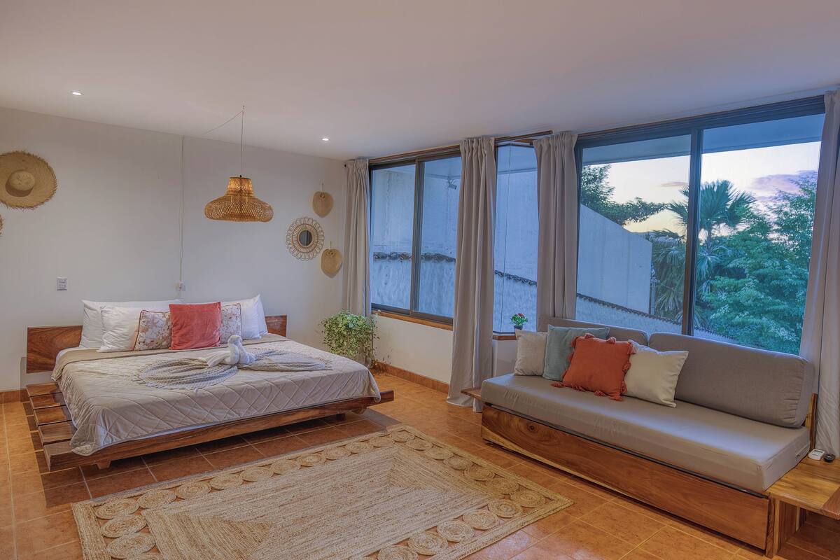 A bedroom with a bed, a sofa, a large window, and a view of greenery outside.
