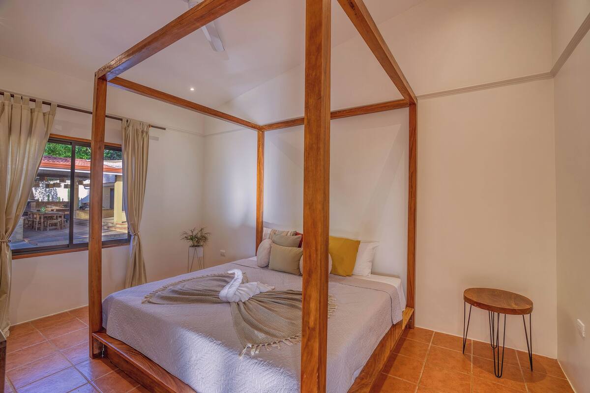 A four-poster bed with a wooden frame, a small round table, and a view of the outside through a window.