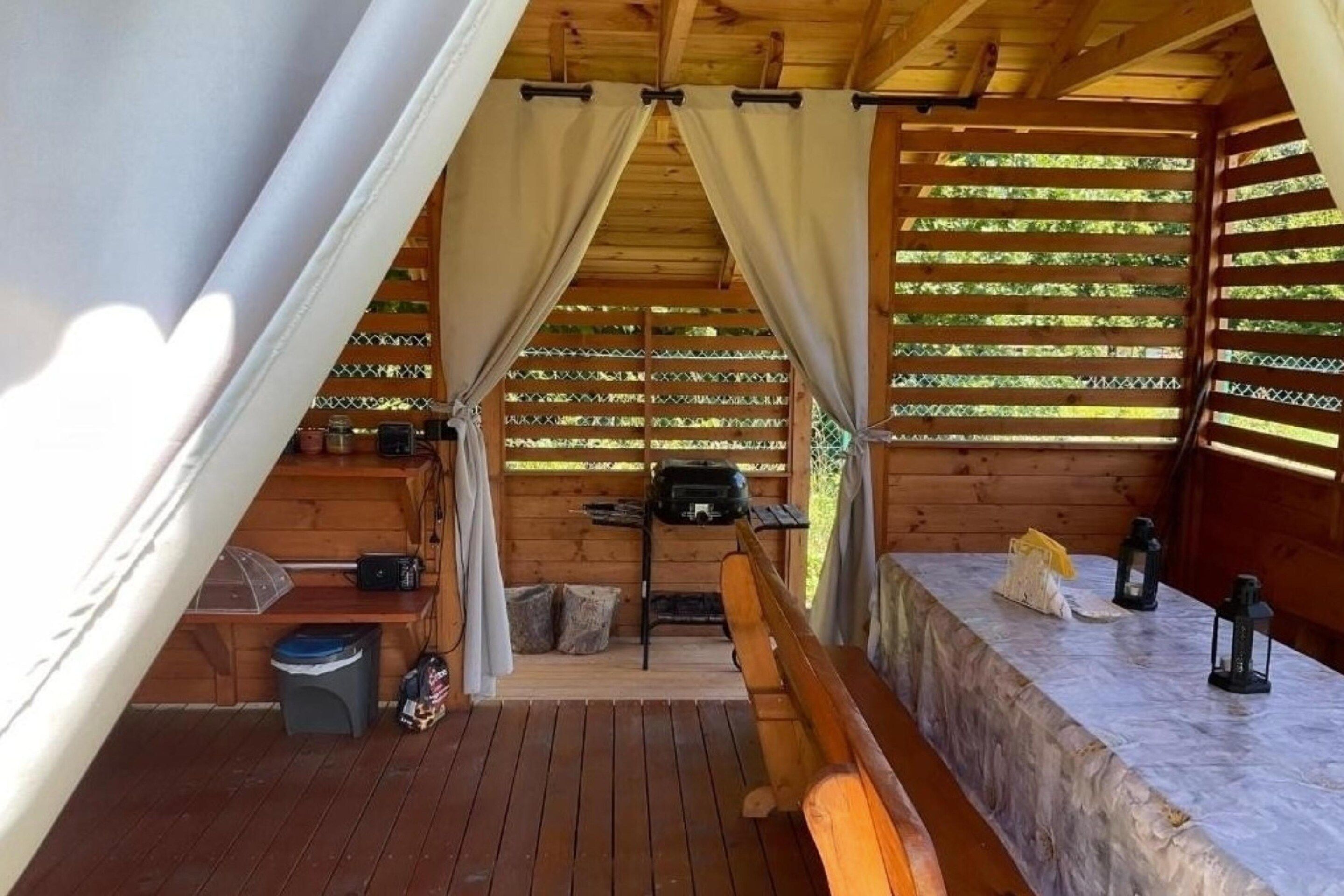 A wooden tent with a table and benches, a grill, and a trash bin.