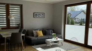 Ein modernes Wohnzimmer mit grauem Sofa, einem Esstisch mit Stühlen, einem weißen Couchtisch und einem großen Fenster mit Jalousien.
