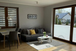 A modern living room with a grey sofa, a dining table with chairs, a white coffee table, and a large window with blinds. - Holiday Home, Large Garden With Playground, Gazebo (Ustka)