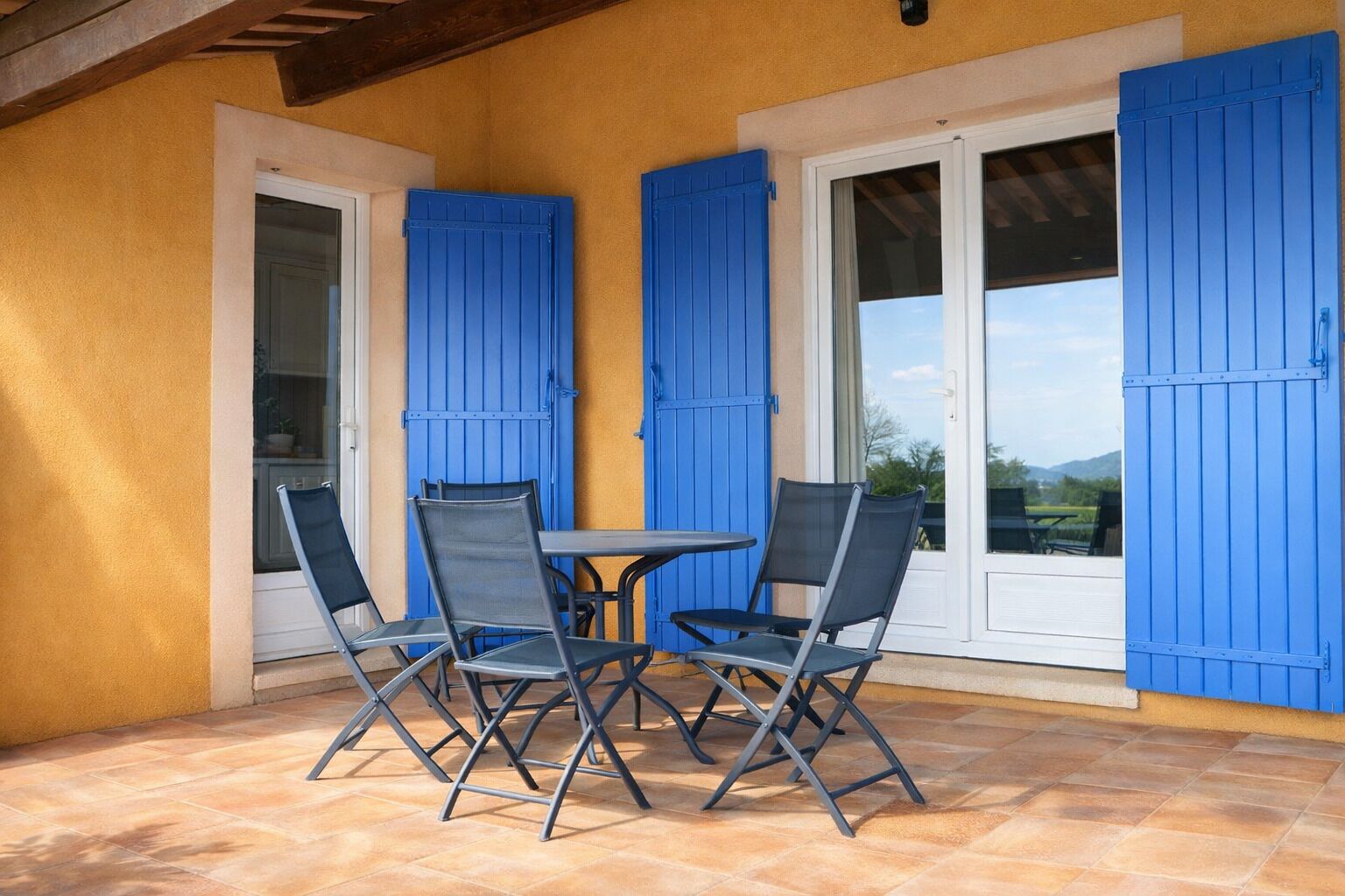 A patio with a table and four chairs, blue shutters, and a view of the outdoors.