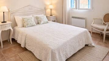 A bedroom with a bed, two bedside tables with lamps, a chair, a window with curtains, and a radiator.