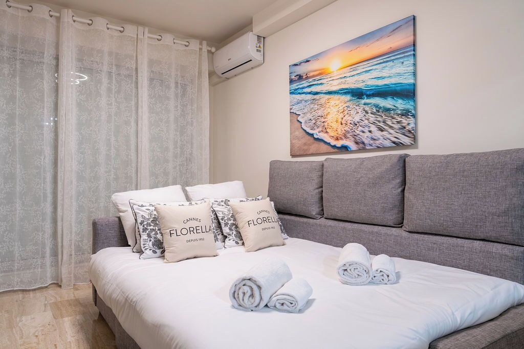 Un dormitorio con cama, cabecera gris, ropa de cama blanca y un cuadro de un atardecer sobre el mar.