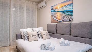 Un dormitorio con cama, cabecera gris, ropa de cama blanca y un cuadro de un atardecer sobre el mar.