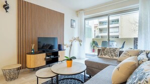 A modern living room with a flat-screen TV, a wooden panel wall, a sofa, a coffee table, and a potted orchid. - Le Roxane joli cocon avec petit terrasse (Cannes)